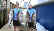 Two smiling men are proudly displaying large fish they appear to have caught, standing on a dockside with a boat in the background.