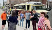 A group of people seem to be listening attentively to a person speaking in front of them, with a trolley bus labeled 