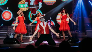 A group of performers in festive red outfits are energetically singing and dancing on stage with a Christmas-themed backdrop.