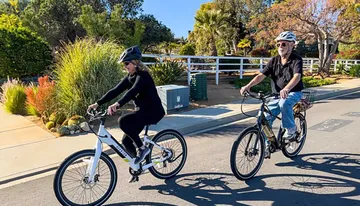 Two individuals are enjoying a sunny day while riding bicycles on a path with lush vegetation around them.