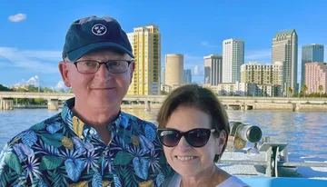 A smiling couple poses in front of a scenic city skyline and a waterway, with clear blue skies overhead.