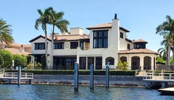 The image shows a large, luxurious waterfront home with palm trees and a private dock under a bright blue sky.