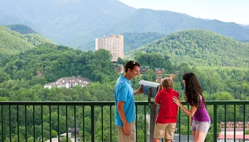 A man and a woman are with a young child who is using a viewing telescope on a balcony overlooking a lush green mountainous landscape with buildings nestled among the trees.