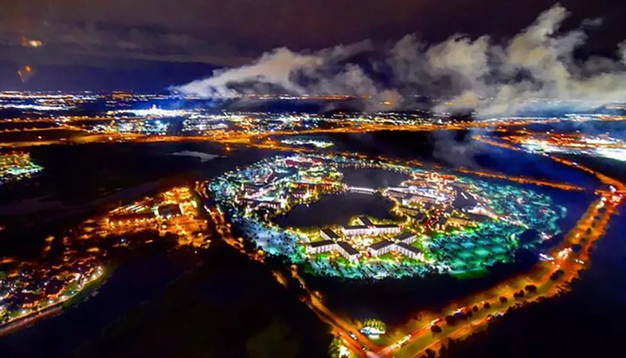 The image shows a vibrant aerial nighttime view of a brightly lit complex with adjacent roads and surrounding dark landscape, under a cloudy sky.