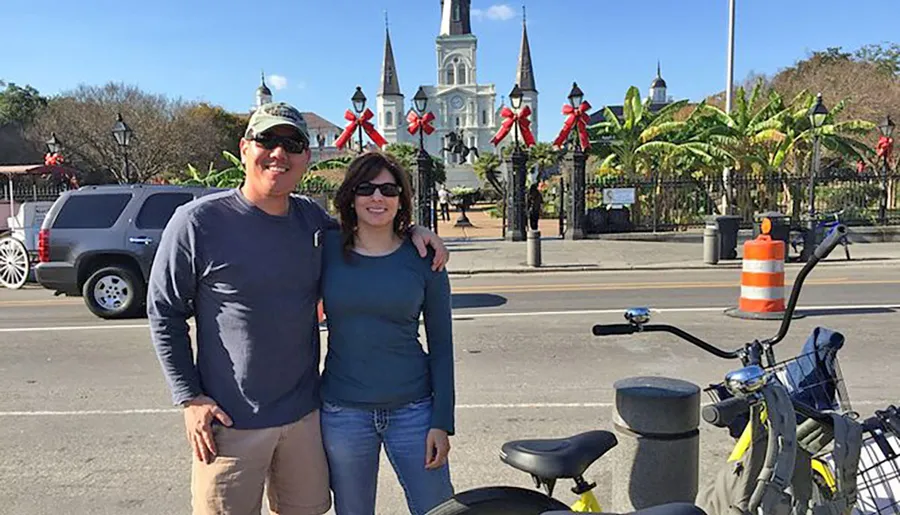 Two people pose for a photo in front of the iconic St. Louis Cathedral in New Orleans, decorated with red bows, under sunny skies.