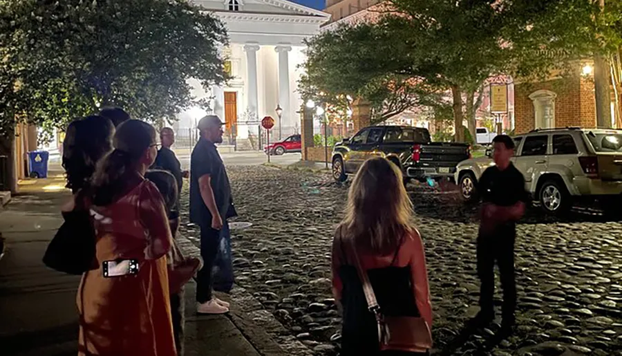 A group of people is standing on a cobblestone street at night, possibly on a tour, with a building featuring white columns in the background.