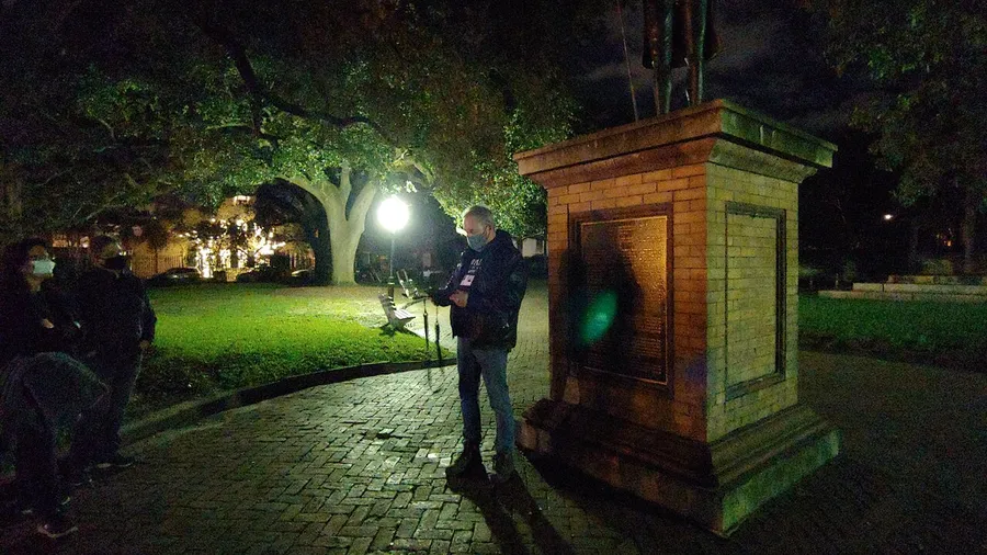 A group of people gather at night in a park illuminated by a lamp, with one person standing and reading from a paper by a monument.