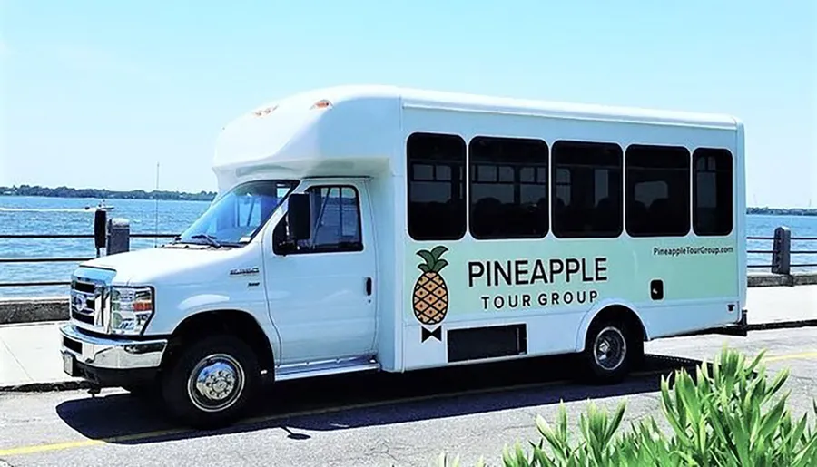 A white shuttle bus labeled PINEAPPLE TOUR GROUP is parked near a body of water on a sunny day.