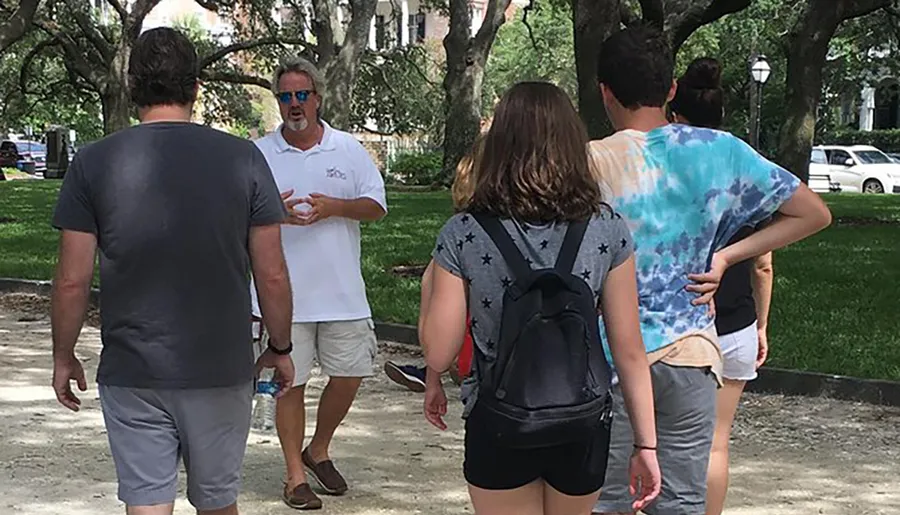 A man is standing and explaining something to a group of attentive listeners in an outdoor setting with greenery in the background.