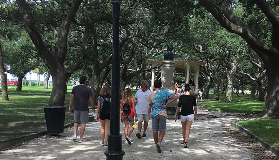 A group of people stroll down a shaded pathway lined by large trees, with a sculpture and gazebo visible in the background.