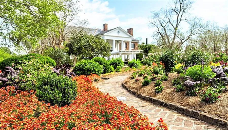 A vibrant garden with colorful flowers leads to a classic white house with a porch.
