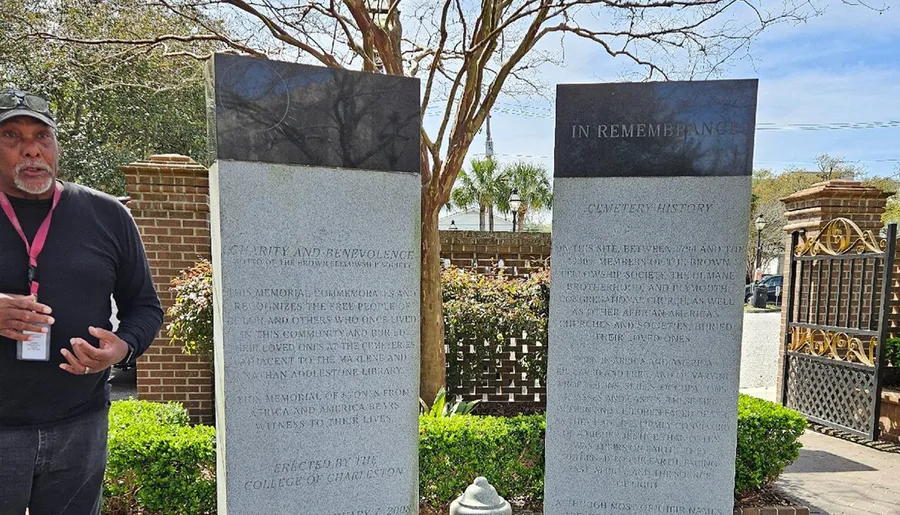 A person is speaking next to memorial stones at the entrance of a cemetery on a sunny day.