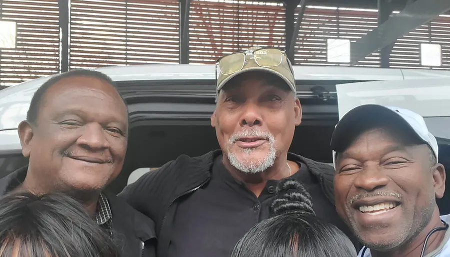 Three men are smiling for a group selfie, with one partially obscured by another's head, and a car and patterned architectural structure in the background.