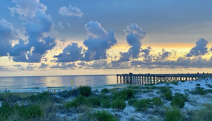 The image captures a tranquil sunset over a beach with a wooden pier extending into the calm sea, under a sky filled with dramatic clouds.