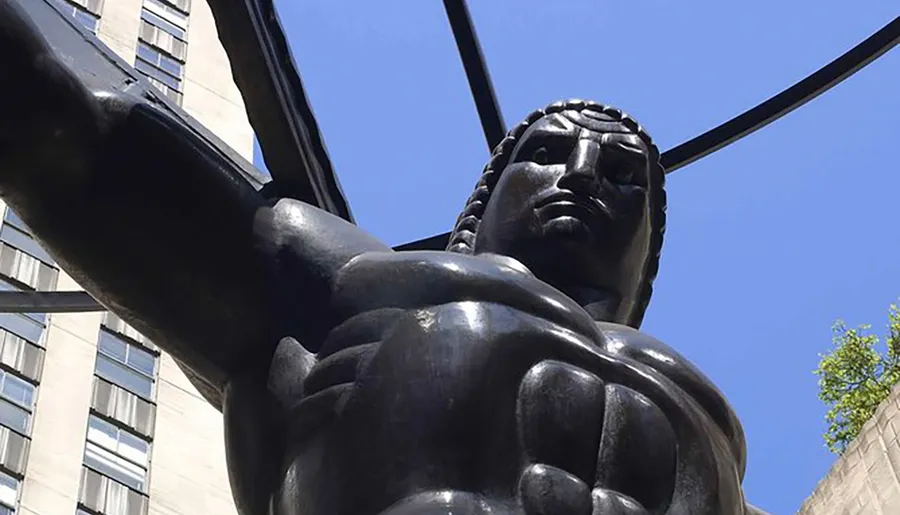 The image captures a view looking up at a large, dark bronze statue of a muscular man with defined features and an outstretched arm against a clear sky.