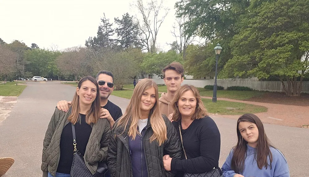 A group of six individuals possibly a family are smiling and posing for a photo outdoors on a path with trees and lampposts in the background