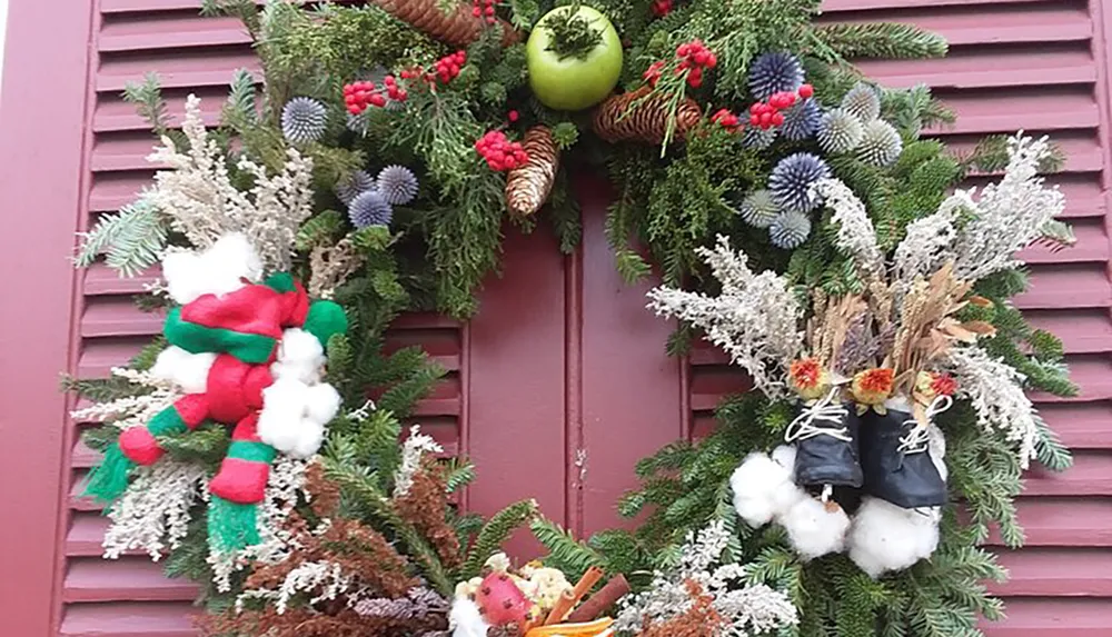 This image features an elaborate and festive Christmas wreath adorned with greenery red berries pine cones a large green and red bow and what appears to be a pair of miniature boots displayed on a pastel burgundy door