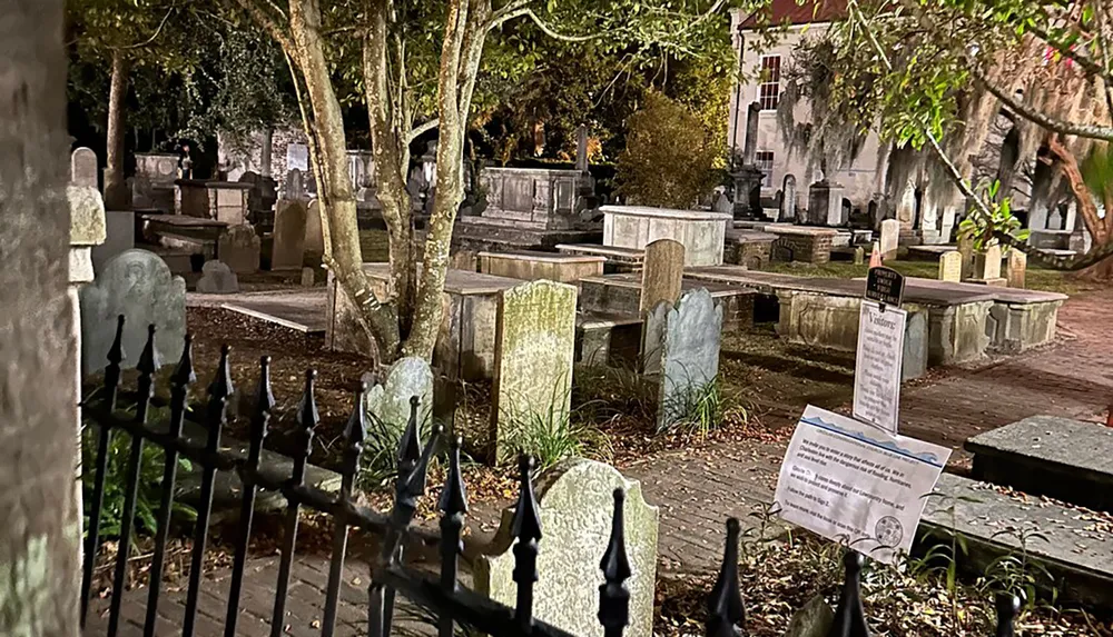 The image shows a historical cemetery at night illuminated by artificial light and enclosed by a wrought iron fence with aged gravestones and monuments scattered among the trees