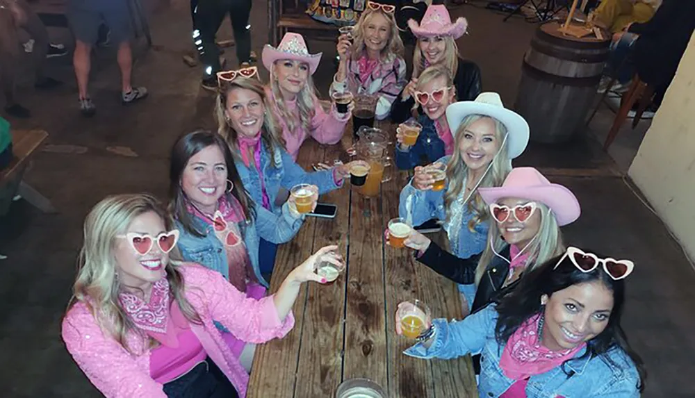 A group of smiling people is having a themed gathering dressed in pink and cowboy hats toasting with drinks at a wooden table