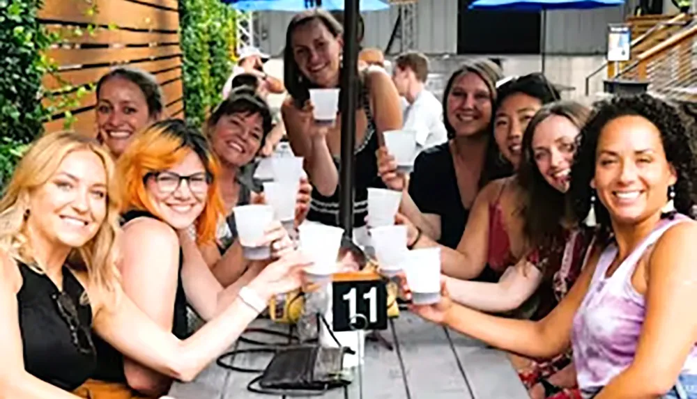 A group of people is smiling at the camera while holding up drinks at an outdoor table marked with the number 11