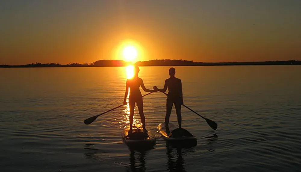Two people are standing on paddleboards on calm water holding paddles and hands against a beautiful sunset backdrop