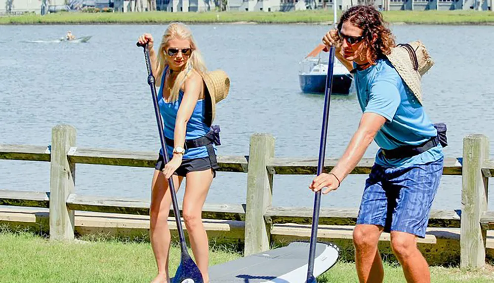 A man and a woman are preparing to go stand-up paddleboarding near a wooden fence by the water