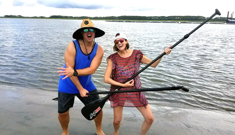 Two people are playfully posing with a stand-up paddleboard paddle near the water appearing happy and energetic