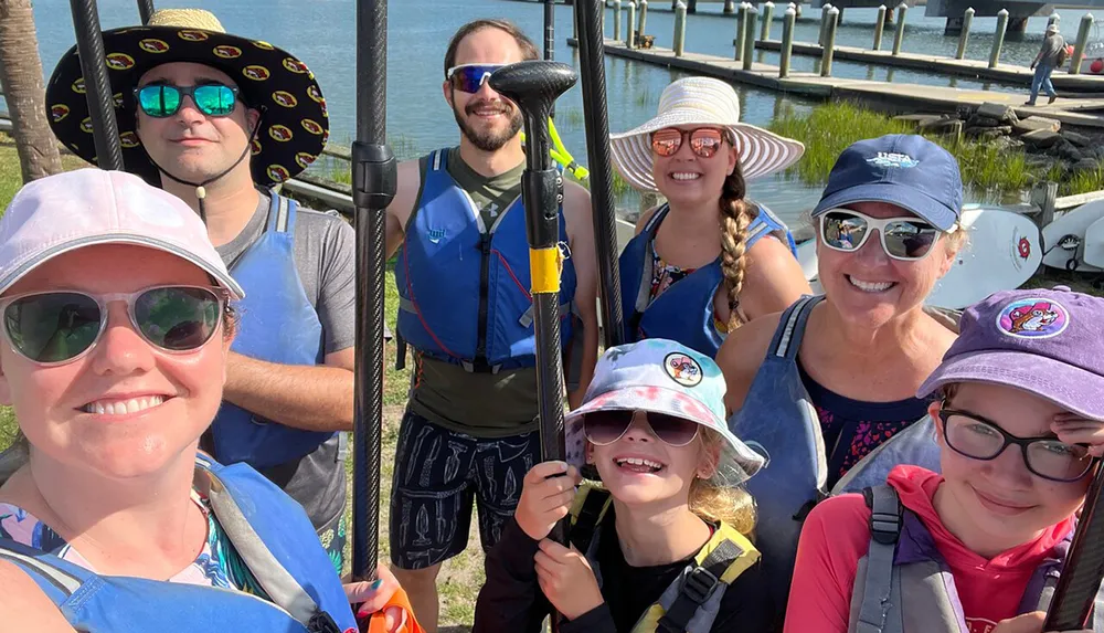 A group of eight smiling people equipped with paddles and wearing life jackets and sun hats appears ready for a kayaking adventure by the water