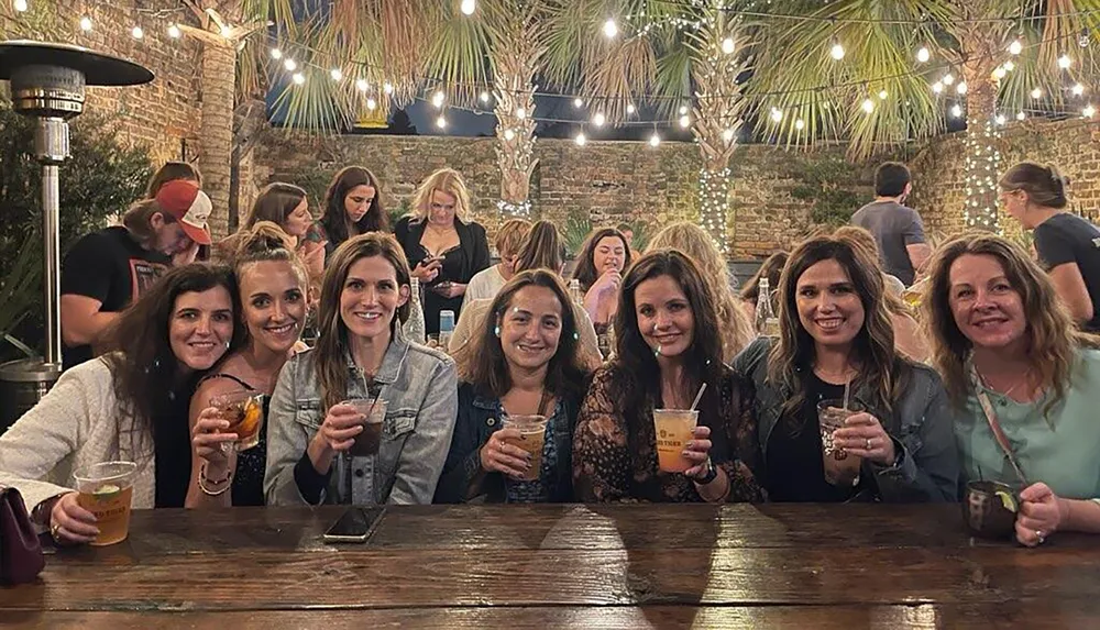 A group of smiling women are gathered at an outdoor table holding drinks with a background of fairy lights and a brick wall