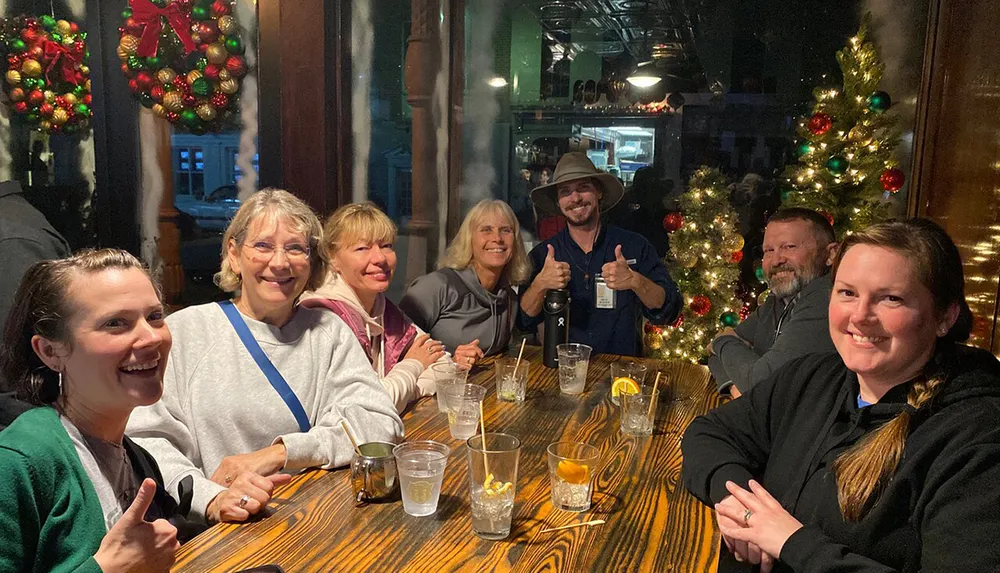 A group of cheerful people are sitting at a long table with drinks giving thumbs-up and smiling in a festive setting with Christmas decorations in the background