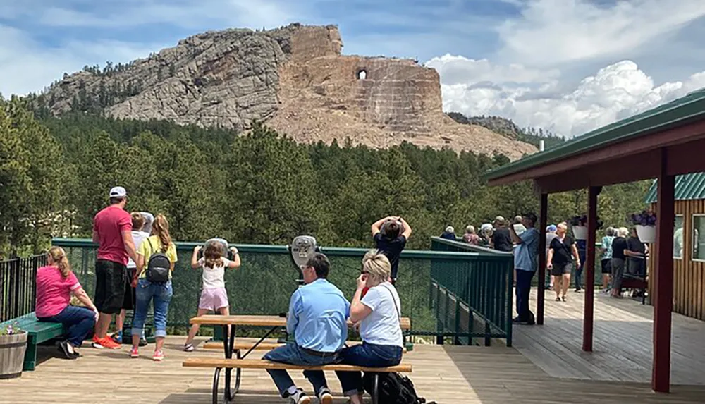 Visitors at an observation deck are looking at and taking photos of a large mountainous monument under a partly cloudy sky