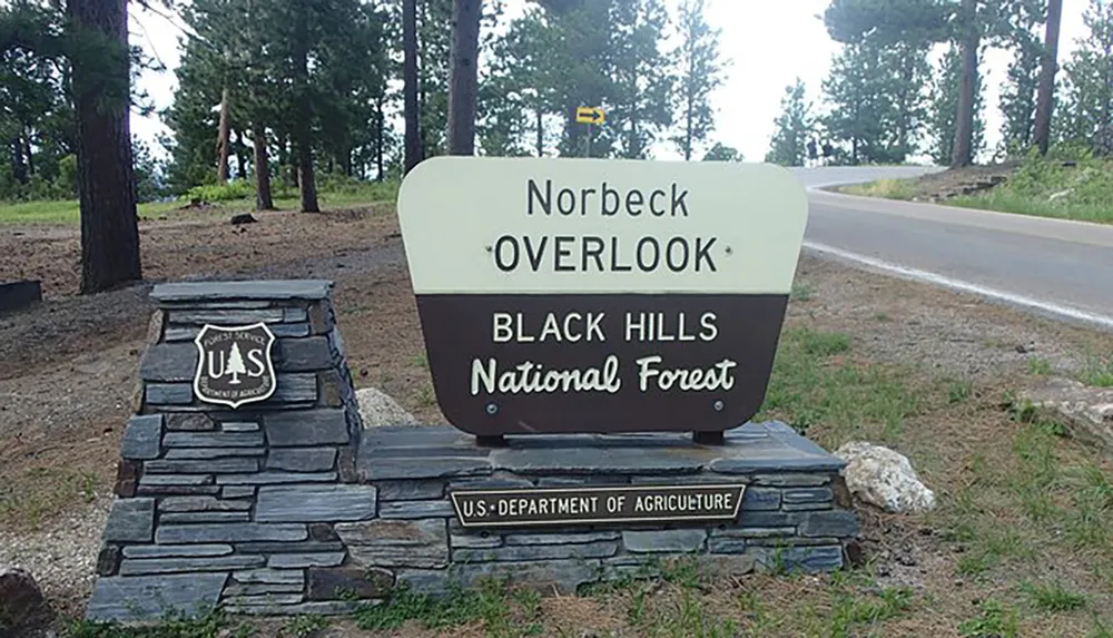 The image shows a sign for Norbeck Overlook in the Black Hills National Forest with a forested background and a road running beside it