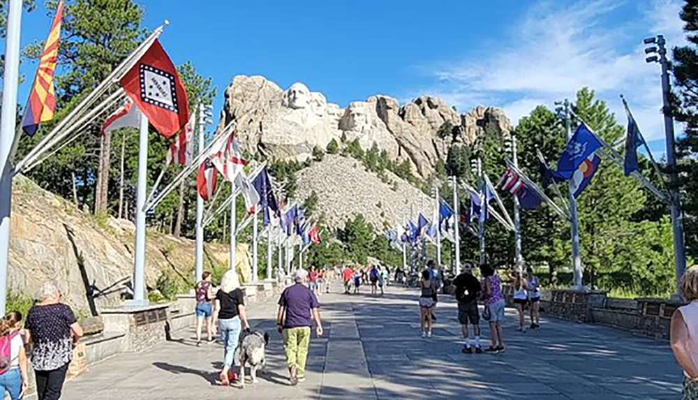 Visitors are walking along the flag-lined avenue that leads to the Mount Rushmore National Memorial with the carved faces of the US presidents visible in the background