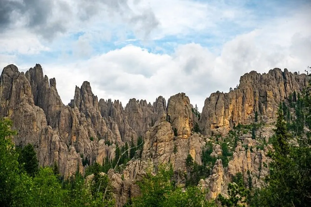 The image shows a rugged and jagged mountain range piercing the sky surrounded by lush green foliage under a cloudy sky
