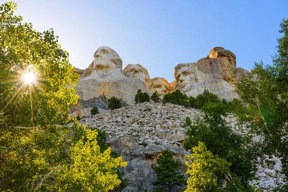 The image captures the sun shining through the trees beside the iconic Mount Rushmore National Memorial which features the carved faces of four former US presidents