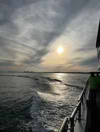 The image captures the view from the side of a boat as it moves across the water, leaving a trail of waves behind, under a partly cloudy sky with the sun hanging low near the horizon.