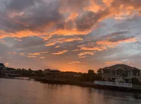 The image captures a breathtaking sunset with vibrant orange and pink hues in the sky, reflecting over a tranquil river beside a neighborhood with elegant waterfront homes.