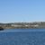 A serene lake stretches across the foreground with a hilly landscape featuring scattered buildings and a water tower under a clear blue sky.