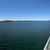 The image shows a panoramic view of a lake from a boat deck with empty chairs and a few people enjoying the sunny weather.