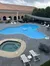 This image shows an outdoor swimming pool area with lounge chairs and a hot tub, situated next to a building with arched openings, all under an overcast sky.