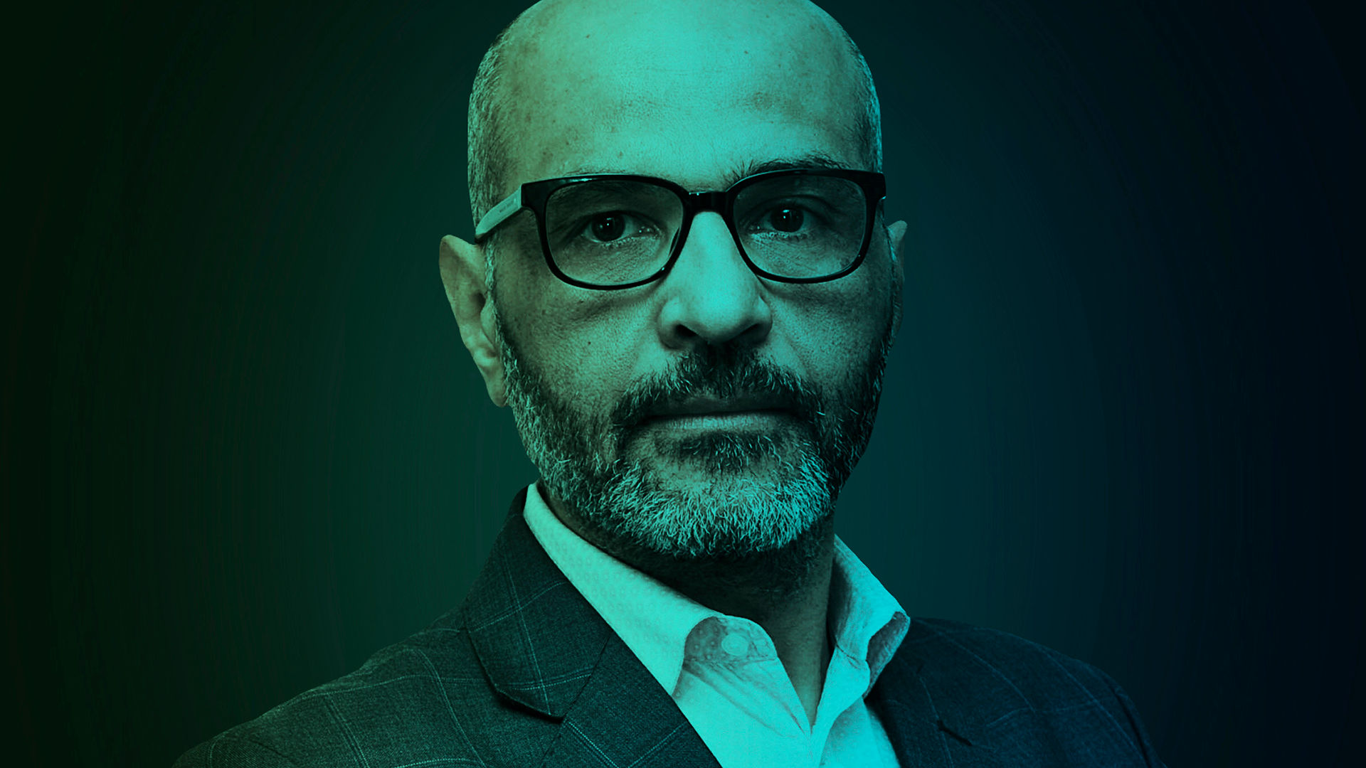 A person with grey hair and a neatly trimmed beard, dressed smartly in a suit and designer glasses.