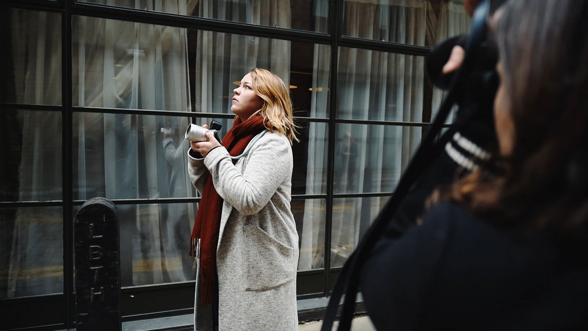 A person with long and blonde hair, wearing a light coloured coat and red scarf, preparing to take a photograph of something in the distance