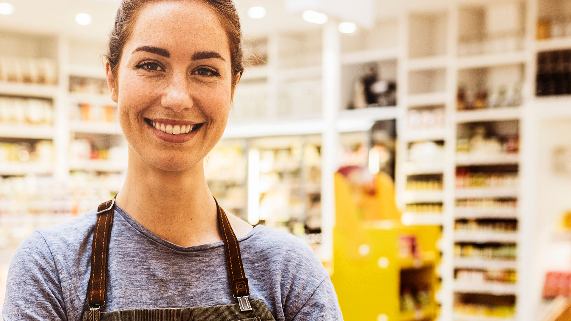 A smiling shop worker wearing a grey shirt and brown apron