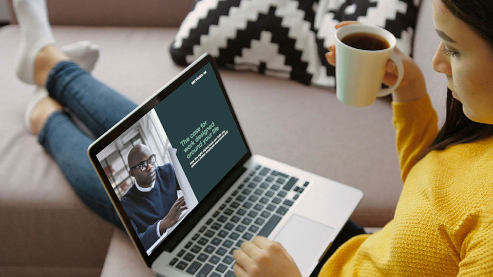 A person sitting down drinking coffee, looking at a laptop with the ServiceNow website on the screen