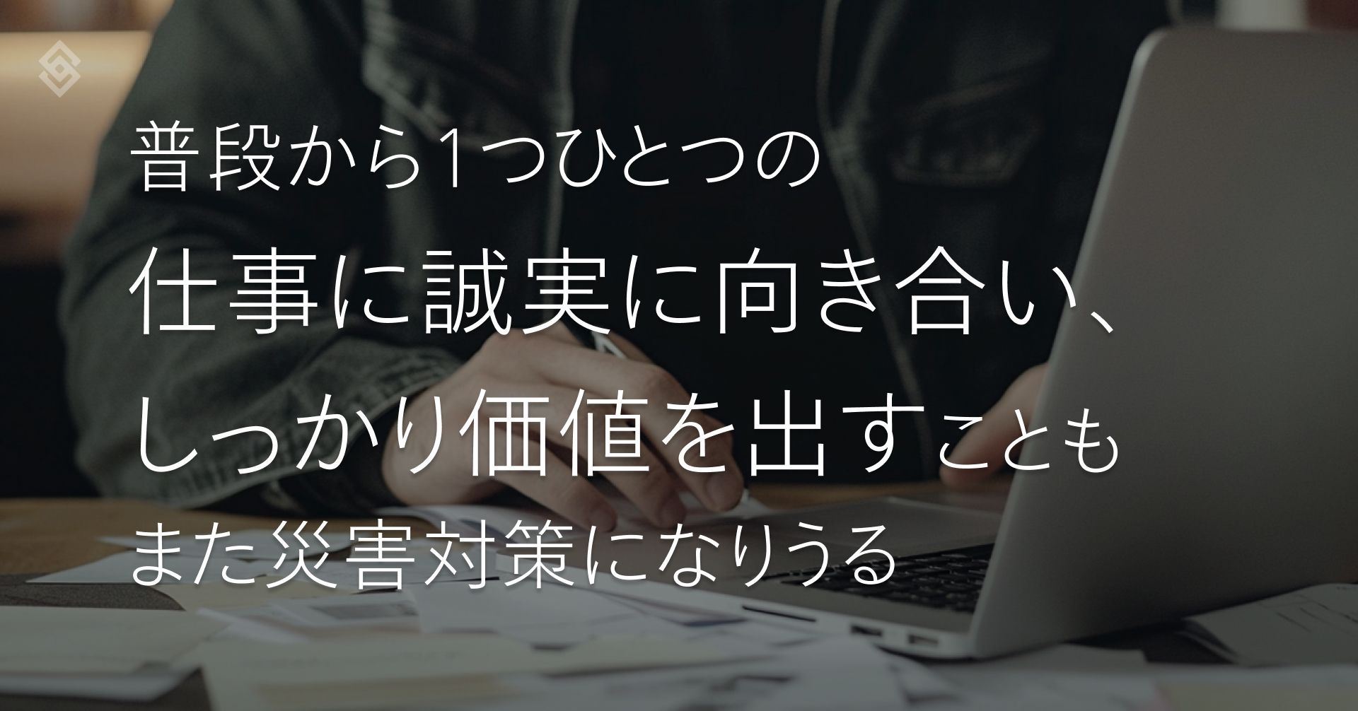 パソコンに向かって作業をしている人。机の上には紙が乱雑に置かれている