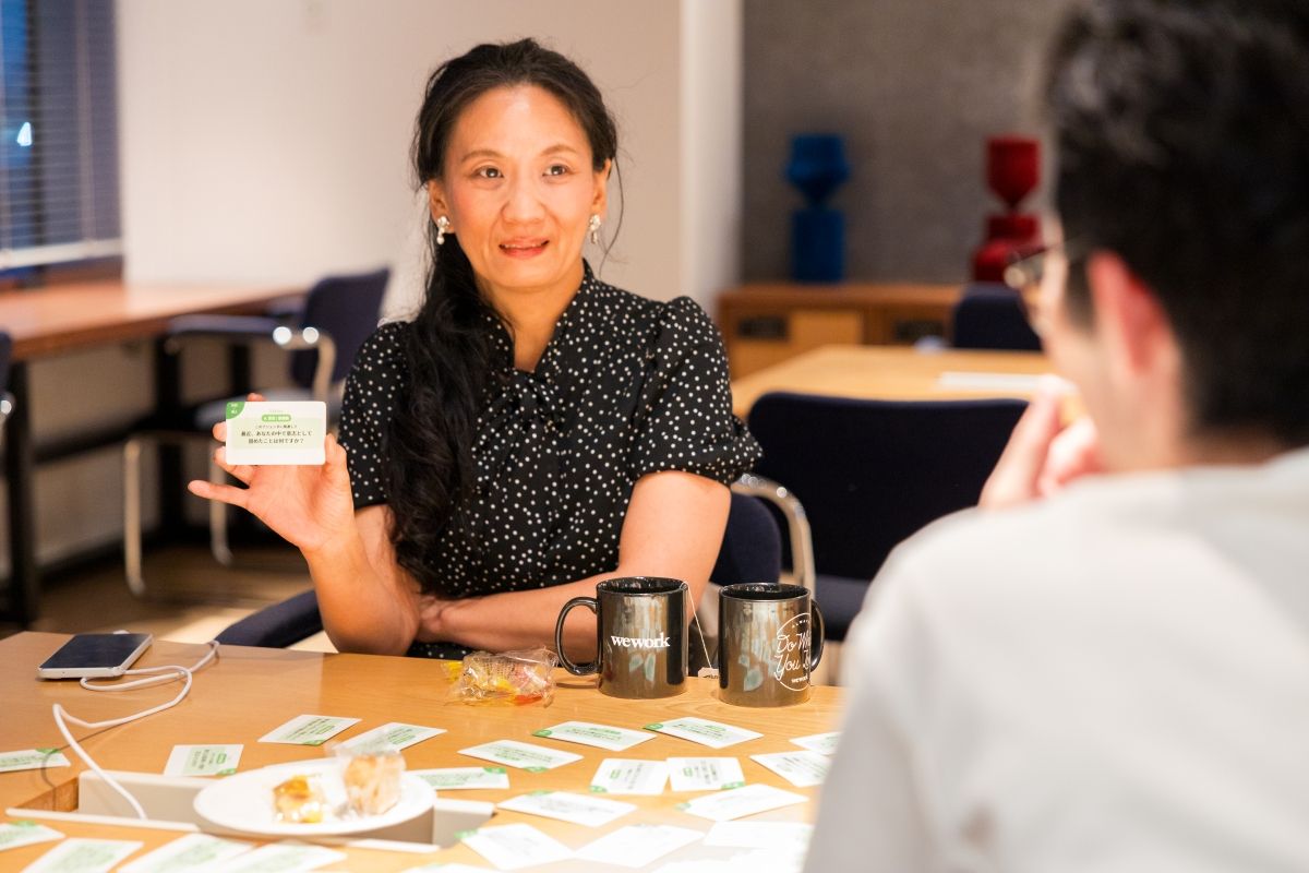 テーブルで向かい合って座る女性が、カードを手に取り説明している様子。机上には複数のカードとマグカップ、軽食が並んでいる。