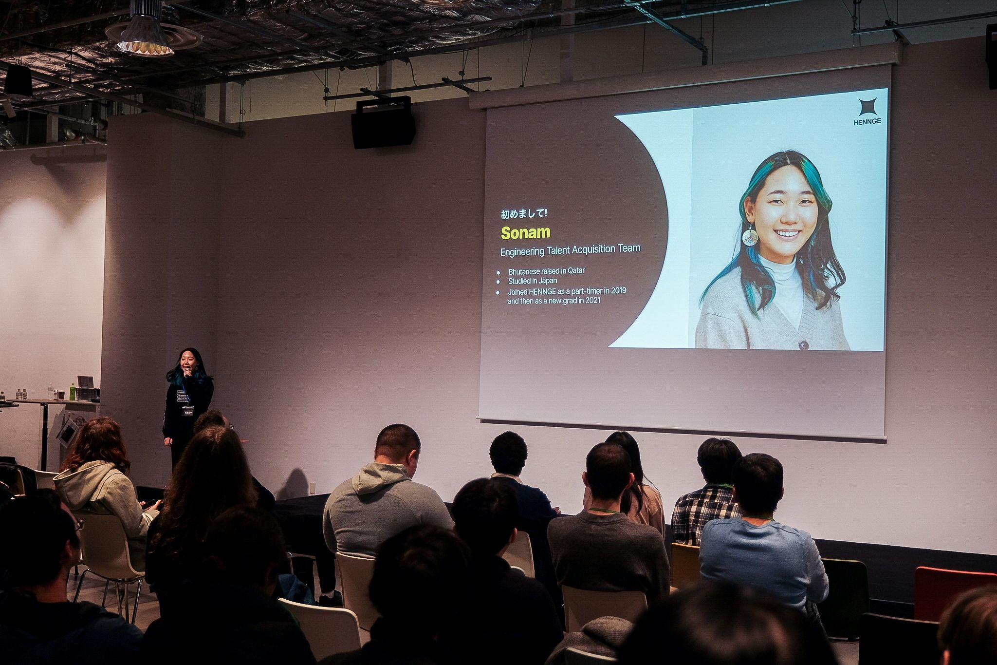 スライドを背景に、HENGEの登壇者が自己紹介を行い、観客が着席して話を聞いている様子。A speaker introducing herself on stage, with a slide projected behind her and the audience listening attentively.