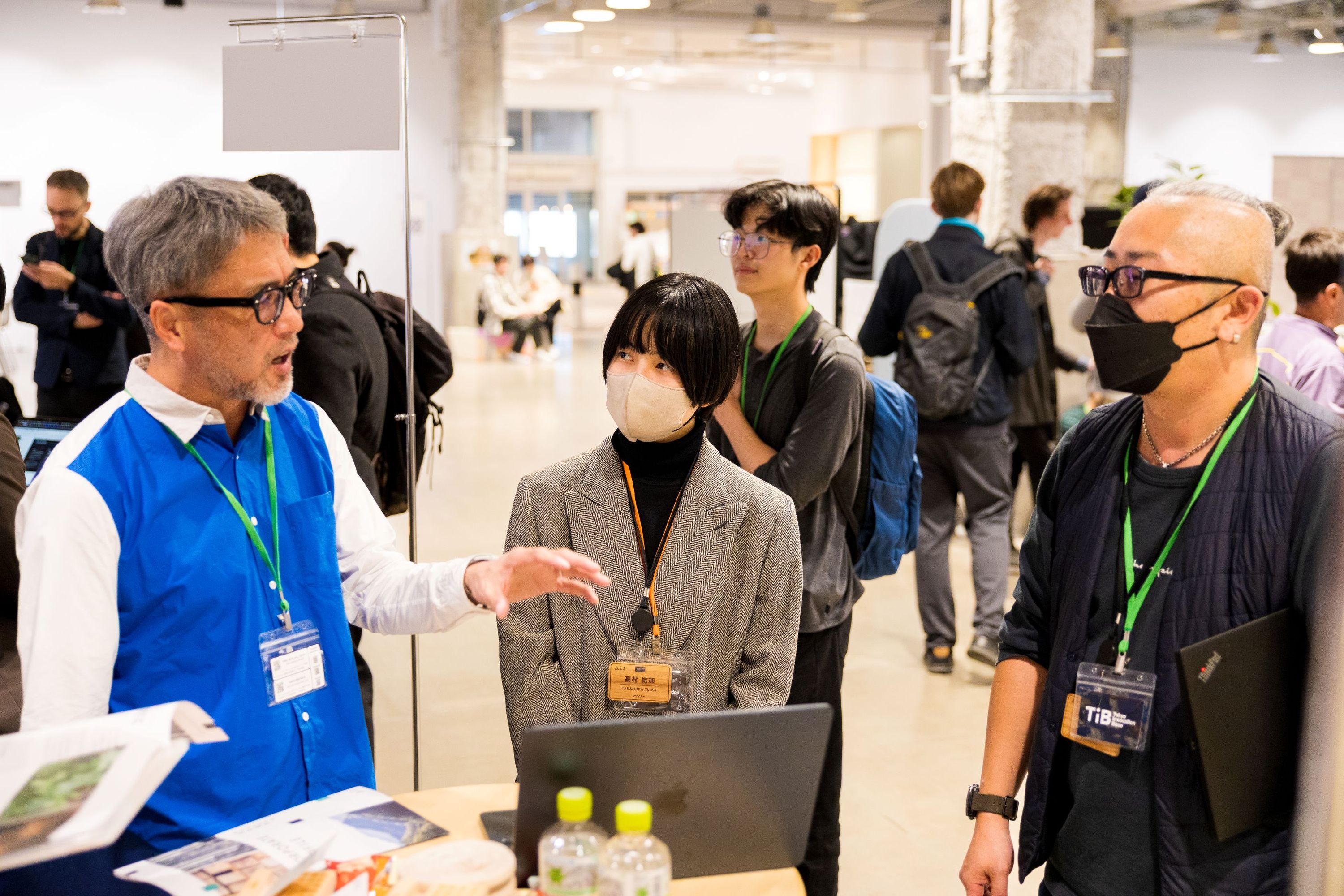 ブースで、担当者がノートパソコンや資料を使いながらプロジェクトについて説明し、参加者が話を聞いている様子。Attendees listening to a representative explaining a project at a booth, with a laptop and printed materials on the table.