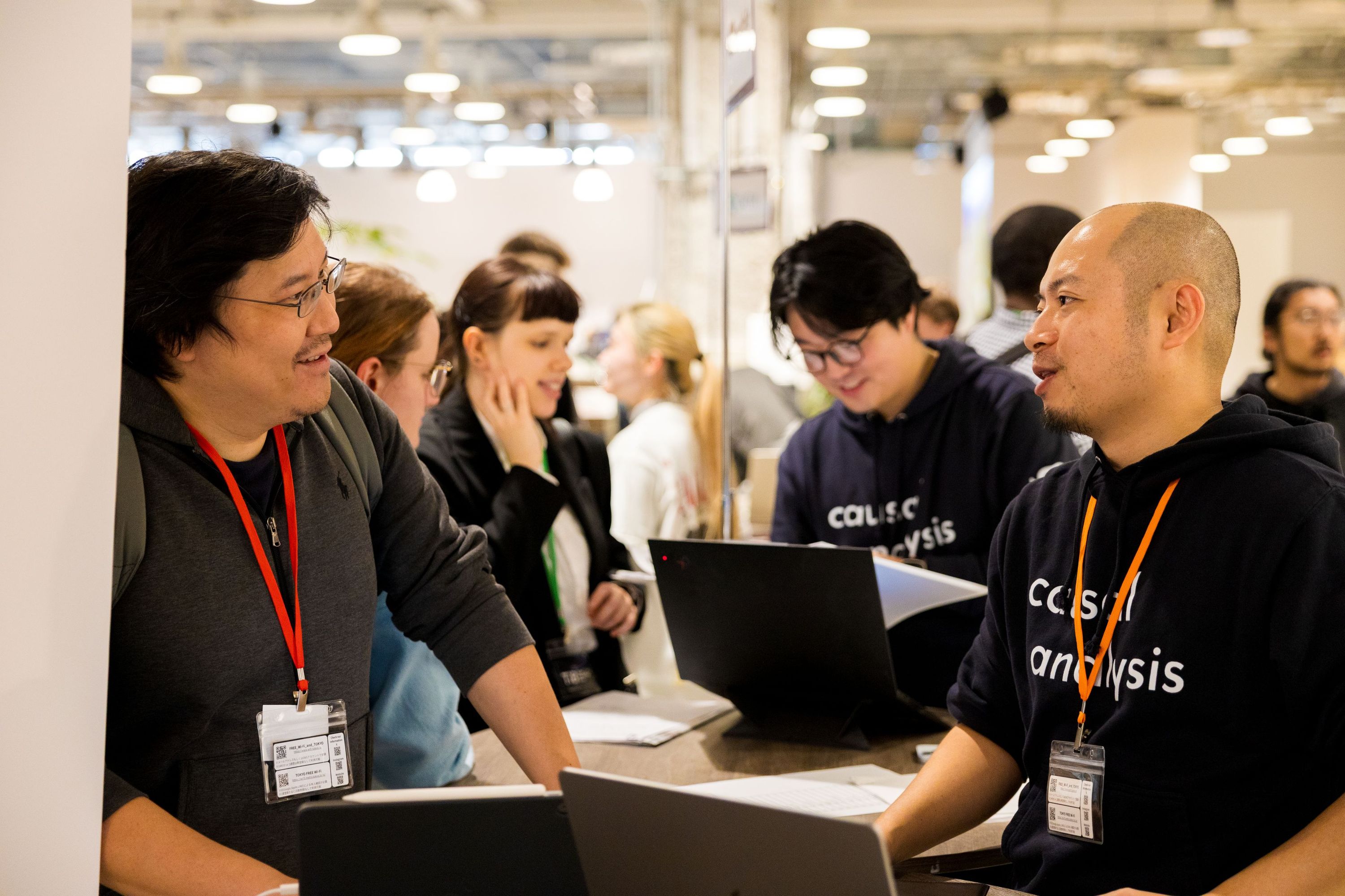 企業ブースで、参加者と担当者がカウンターを囲み、キャリアや業務内容について話している様子。Attendees engaging in conversation with company representatives at a booth, discussing opportunities while standing around a counter.