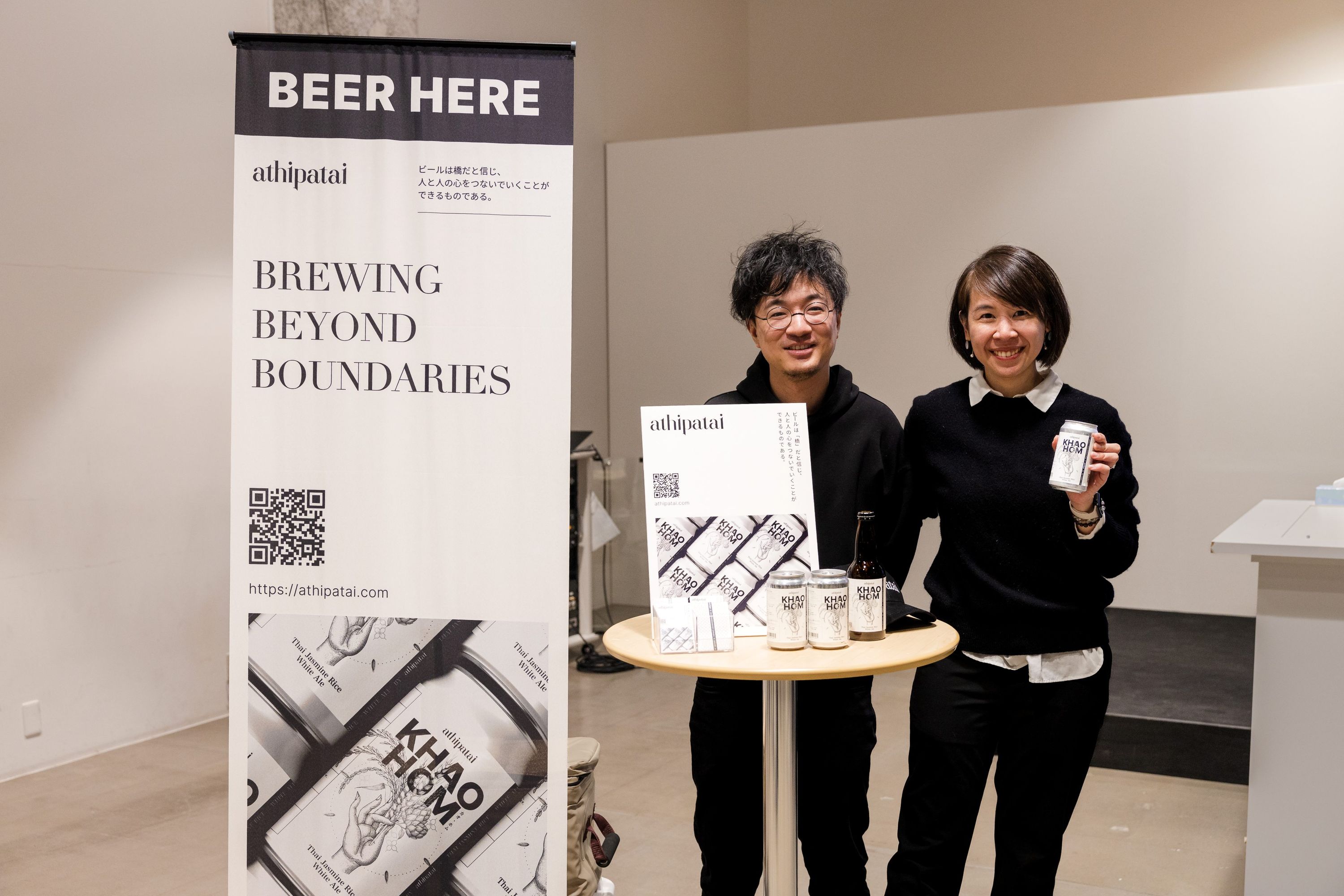 クラフトビールブランド「athipatai」の展示ブースで、出展者2名がビール缶を手に立ち、背景には「BEER HERE」「BREWING BEYOND BOUNDARIES」と書かれたバナーが設置されている。Two exhibitors standing beside a display for athipatai beer, holding cans and standing next to a banner that reads “BEER HERE” and “BREWING BEYOND BOUNDARIES.”